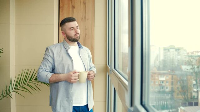 bearded handsome man stands by window with a cup in his hand on the balcony and drinks a hot coffee or tea. male in casual clothes enjoys view through in office or home, relaxing or timeout at work