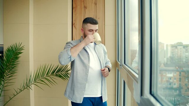 bearded handsome man stands by window with a cup in his hand on the balcony and drinks a hot coffee or tea. male in casual clothes enjoys view through in office or home, relaxing or timeout at work