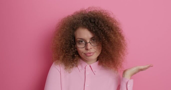 People human perception reaction concept. Hesitant curly haired woman spreads palms feels doubtful isolated over pink background. Indecisive clueless female shrugs shoulders with perplexed expression