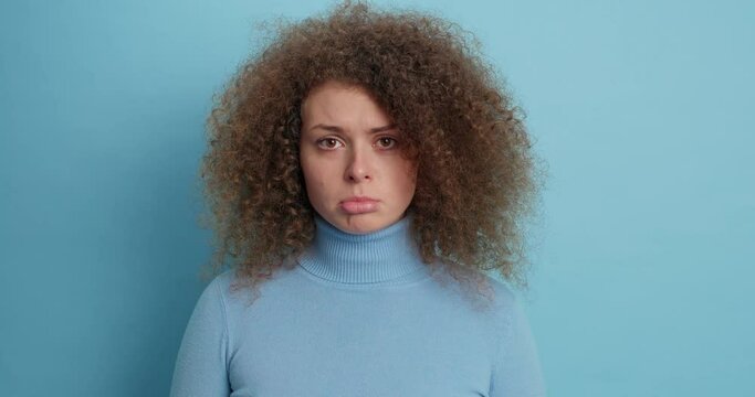 Upset gloomy curly haired Caucasian woman looks sadly at camera has sad expression purses lower lip wears turtleneck isolated over blue background complains as hears something disappointing.