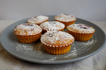 close up six round pumpkin muffins sprinkled with powdered sugar lie on a grey round plate on a beige tablecloth side view . homemade cakes