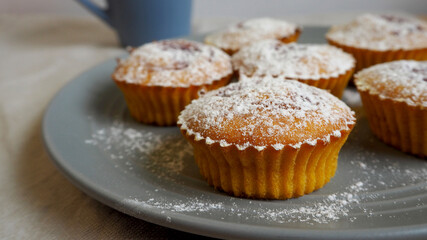 several pumpkin muffins sprinkled with powdered sugar in a gray round plate stand on a beige linen tablecloth against the background of a blue mug side view