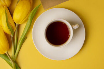 Still life with yellow tulips and a mug of tea with an envelope on a yellow background