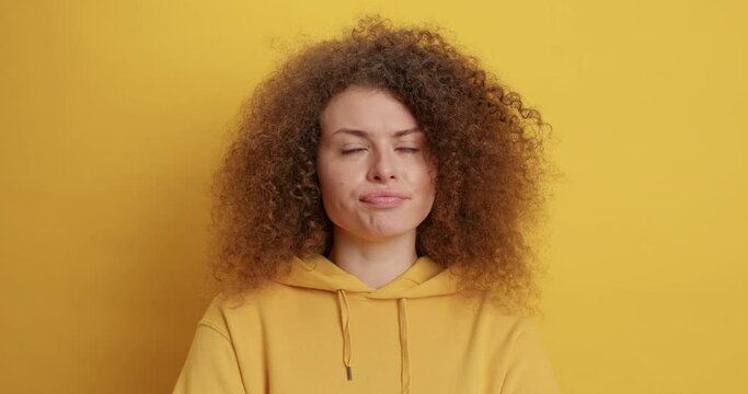 Thoughtful hesitant curly haired European woman has doubts hesitates about deal stands pensive holds chin wears casual sweatshirt isolated over yellow background weighs up choice poses indoor