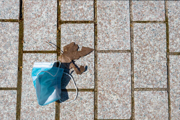 overhead view of a surgical mask lying on the ground next to a dry brown tree leaf. On a pattern of bricks on the ground