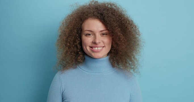 Pretty serious young European woman with curly bushy hair has concentrated look at camera smiles happily dressed in casual turtleneck isolated over blue background. People human emotions concept