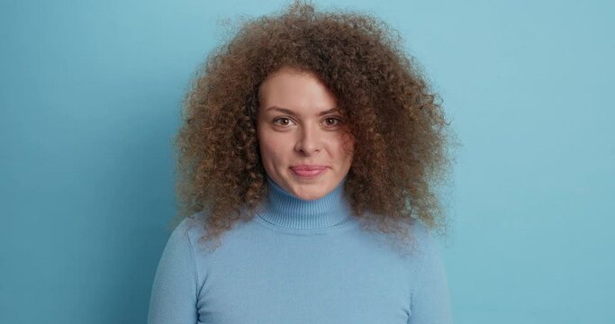 Beautiful curly haired woman licks lips sees something appetizing has tempation to eat sweet dessert smiles gladfully dressed in casual turtleneck isolated over blue background. Mmm how tasty it is