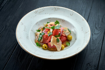 Appetizing, dietary salad with tomatoes, onions, olives and grilled trout in a white plate on a dark wooden background. Healthy food