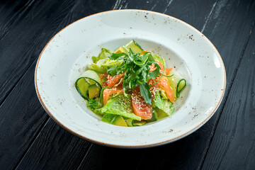 Appetizing, dietary salad with lettuce, cucumber, avocado and lightly salted salmon in a white plate on a dark wood background. Healthy food