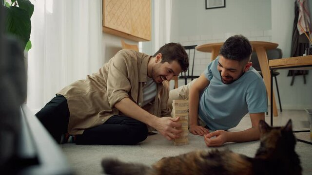 Happy Gay Couple Playing Wooden Block Tower Game while Resting on the Floor. Cheerful Young Boyfriends Enjoy Spending Time Together Having Fun, Playing Games, Talking, Laughing. Pet Cat resting beside