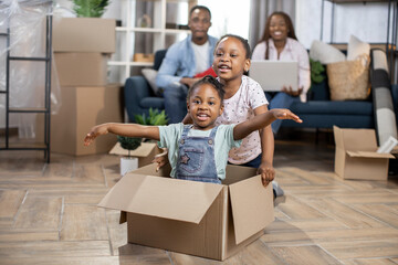 Two little african girls kids playing with boxes while parents relaxing on sofa at modern appartment
