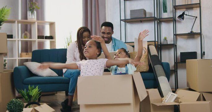 Likable happy smiling modern african american couple sitting on the sofa among carton boxes on moving day while their two small daughters sitting in carton box and depicting the wings of an airplane