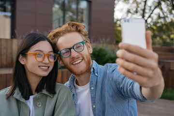 Happy smiling friends influencers using mobile phone, recording video outdoors. Young attractive asian woman and red haired caucasian man wearing stylish eyeglasses taking selfie on the street