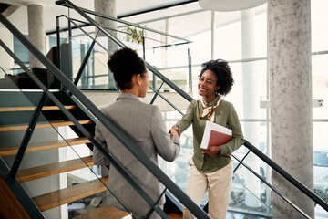 Young happy businesswoman shakes hands with her mentor at they greet each other at staircase in hallway of office building. - obrazy, fototapety, plakaty