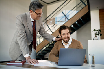 Happy mature businessman encourages his trainee who works on computer at corporate office.