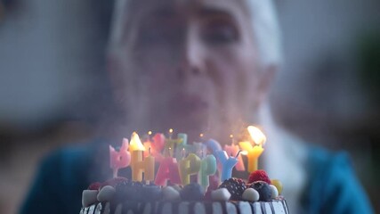 Close-up of festive cake decorated with fresh berries and colored burning candles, blurred on background cheerful aged gray-haired woman blowing out lights during Birthday celebration
