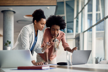 Young black business apprentice talks with her mentor who reviews her work on laptop during meeting in the office.
