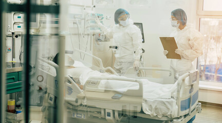 Doctors in protective suits and masks are examining the infected coronavirus young man patient in the hospital ward