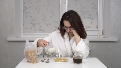 Sleepy young woman in a white robe eats cereals and drinks coffee. Young girl with glasses at home in the morning kitchen.