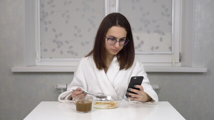 A cheerful young woman in a white coat eats cereals and drinks coffee and uses a smartphone. Young girl with glasses at home in the morning kitchen.