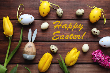 Fototapeta na wymiar Easter card with yellow tulips, wooden bunny, decorative colored and chocolate eggs, and the inscription Happy Easter on a rustic wooden background. Top view, flat lay