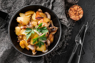 Homemade fried potatoes with mushrooms in a roasting pan with parsley, over a dark background, top view