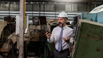 Naklejka na ściany i meble A beard mechanical engineer, supervisor or factory manager is communicating with his staff by using radio or handhold walkie talkie in a warehouse. Smart manager with helmet is getting mad in mission