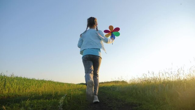 Happy childhood. Cheerful girl are running in the meadow with wind turbines as a sign of freedom. Kid girl holding wind toy. Child plays with windmill. Happy family concept.