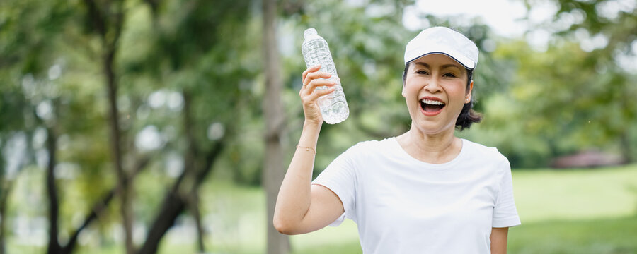 Asian senior woman holding and drinking water in bottle after exercise in garden, relax and exercise concept. Banner image size.