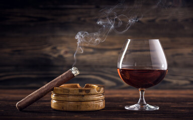 Cognac and cigar. Glass of cognac or brandy with cigars from Cuba Havana and ashtray on natural wooden background. Glass of whiskey with smoke cigar. Alcohol drink on Bar counter in the restaurant.  