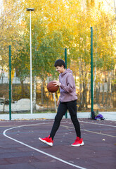 Cute Teenager with orange basketball ball plays basketball on street playground in spring summer. Hobby, active lifestyle, sports activity for kids.	