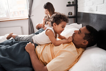 Obraz premium Multiracial parents and cute small kids playing while lying on bed together