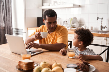 Man drinking tea and chatting with his son while working at the laptop