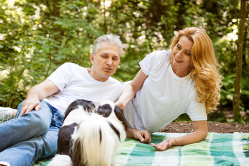 happy mature married couple in forest lie on blanket with dog, picnic concept