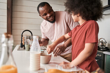 Father in casual clothes and his curly son preparing breakfast at the kitchen