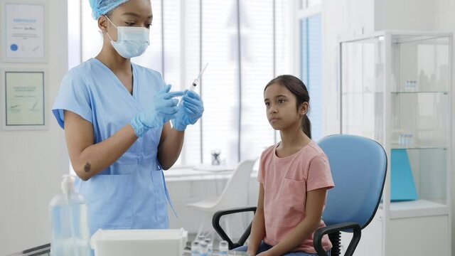 Female medical nurse in protective mask, hat and safety gloves filling syringe and prepering vaccine for young girl. Kid waiting for vaccination while sitting on chair in hospital.