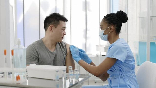 Asian patient getting a shot at the hospital or clinic. African-Amercian doctor in medical face mask injecting young man with flu or Covid-19 antiviral vaccine during mass vaccination campaign.