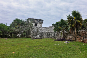 Ruins of the ancient Mayan city of Tulum. A dilapidated stone building stands on the green grass. Around - tropical vegetation. Cloudy sky. Mexico