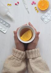 Medicines, pills, thermometer, traditional remedies for colds and flu, women's hands with a cup of tea, on a white wooden background.