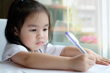 A little asian girl writing an alphabet on a paper. She sitting near the glass window and sun rays shining through it into room.