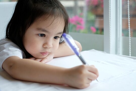 A little asian girl writing an alphabet on a paper. She sitting near the glass window and she looks bored.
