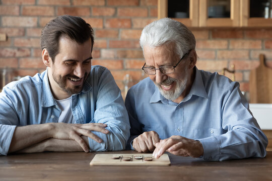 Happy young Caucasian man and senior 60s father have fun engaged in funny interesting logic wooden board game together. Smiling older dad and adult grownup son have fun play checkers draughts.