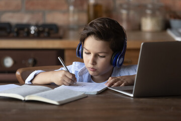 Smart little Caucasian boy in headphones sit at desk at home study online on computer write in notebook. Focused small kid child in earphones have webcam distant lesson on laptop. Education concept. - Powered by Adobe