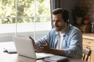 Young Caucasian man wear headphones look at laptop screen talk speak on video call online. Millennial male in earphones use computer have webcam digital virtual conference. Technology concept.