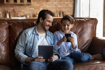 Smiling young 30s Caucasian dad and little 7s son relax on couch at home use modern electronic gadgets together. Happy father and small boy child have fun with tablet smartphone. Technology concept.