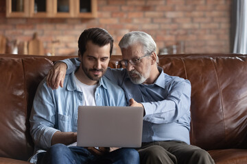 Mature 60s Caucasian dad and millennial adult man kid relax hug on sofa at home using computer...