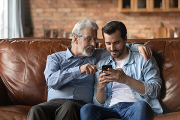 Mature Caucasian father and adult son relax on sofa at home using modern smartphone gadget. Old 60s...