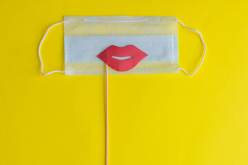 Medical face mask, paper pink lips on stick on bright yellow background, close-up. Celebrating April Fools ' Day during coronavirus pandemic concept.