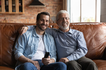 Happy elderly Caucasian grey-haired father and adult 20s son sit relax on couch at home watch TV...