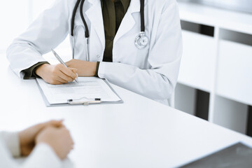 Fototapeta premium Unknown woman-doctor and female patient sitting and talking at medical examination in clinic, close-up. Therapist wearing green blouse is filling up medication history record. Medicine concept